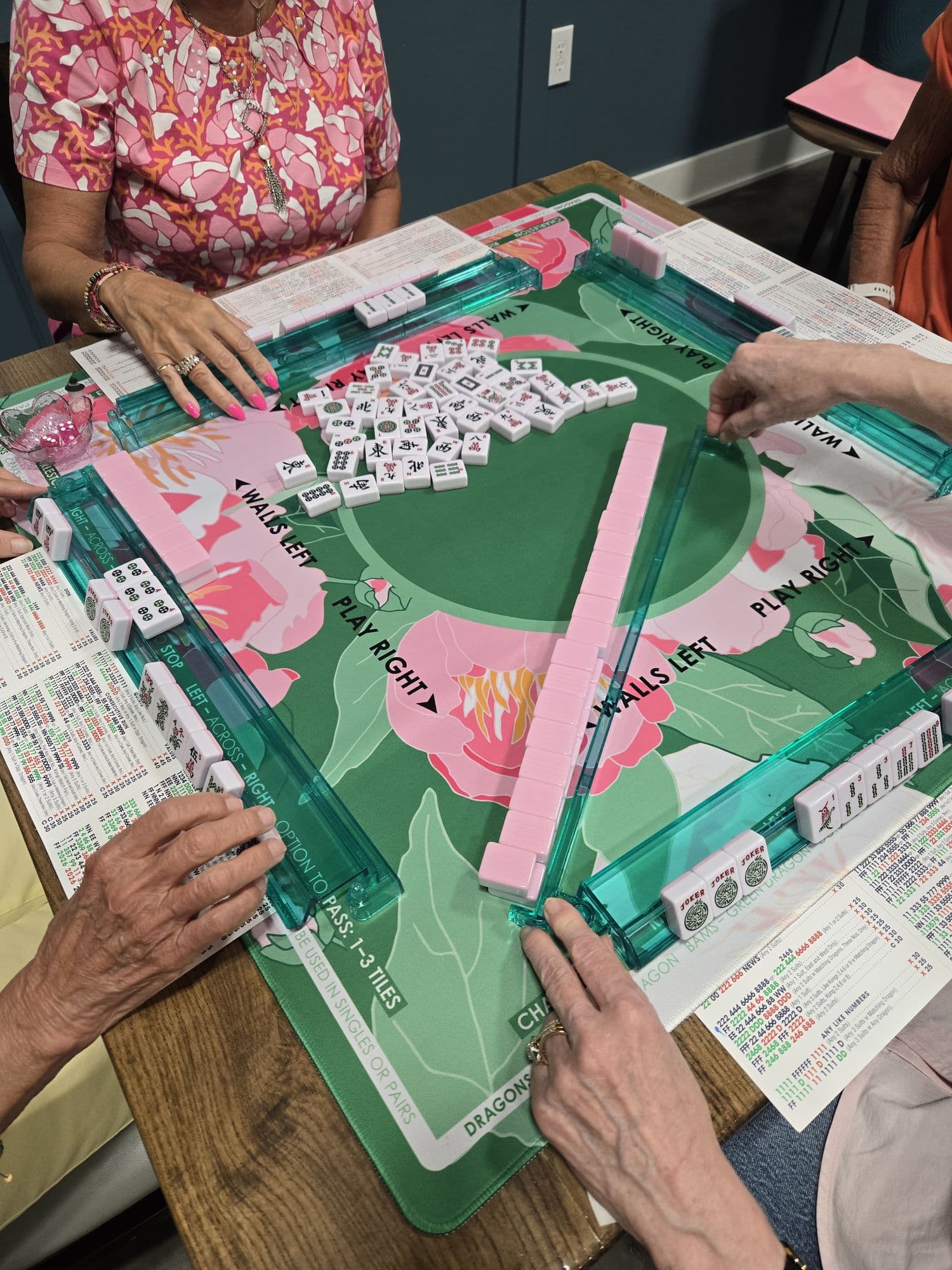 Overhead view of players' hands arranging mahjong tiles on a pink and green table